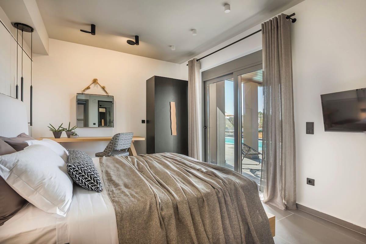 A modern bedroom with a large bed, a flat-screen TV, a mirror, and a sliding glass door to a balcony.