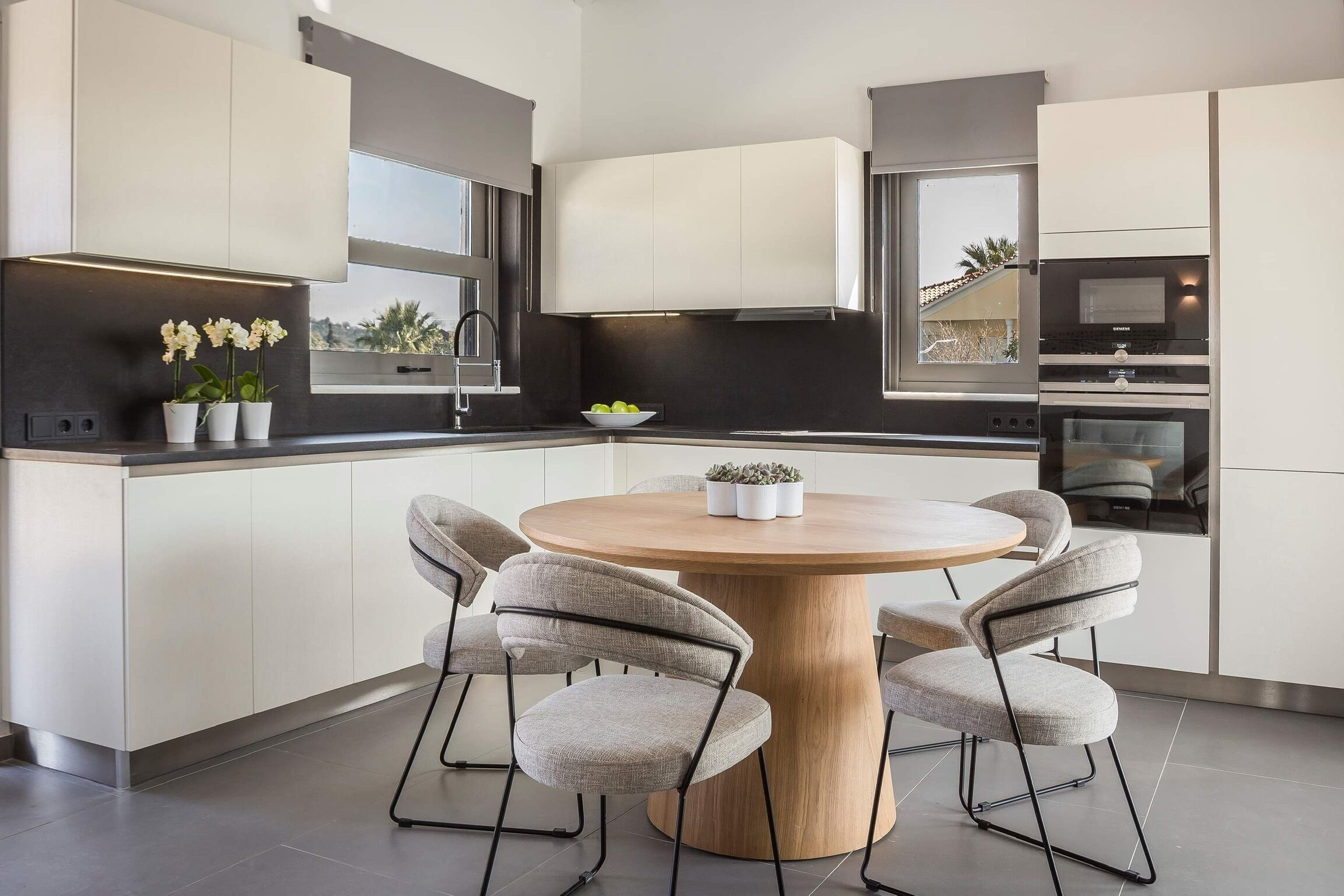 Una cocina moderna con armarios blancos, una mesa redonda de madera y sillas tapizadas en gris.