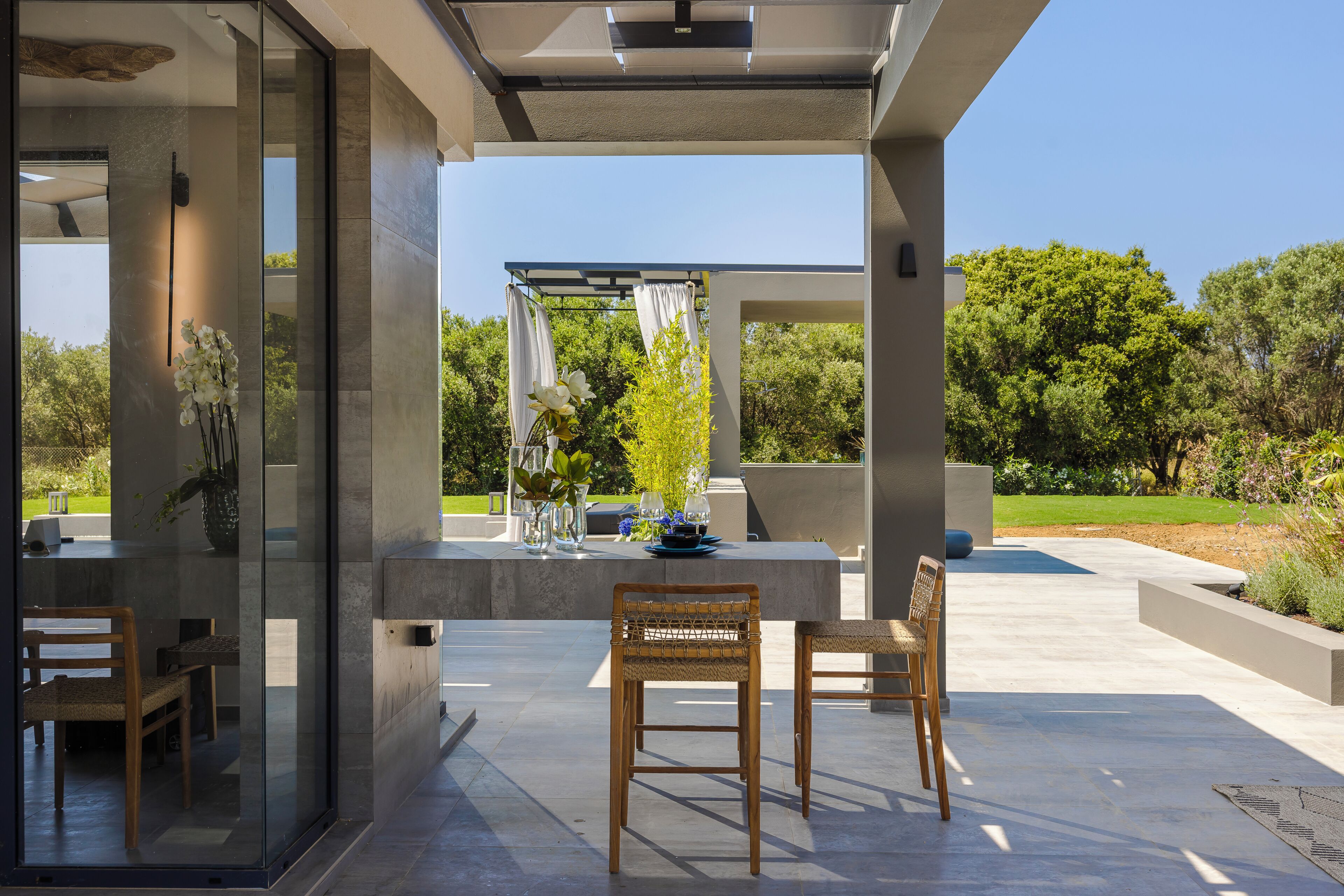 A modern outdoor patio with glass walls, two wicker chairs, a table with a vase, and a pergola with white curtains.