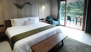 A hotel room with a large bed, a bench, a chair, a table, and a balcony with a view of greenery. - Celebration Land by Miros (Karjat)
