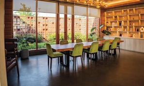 Un comedor con una mesa larga y sillas, ventanas amplias con vistas a zonas verdes, y una estantería con diversos objetos.