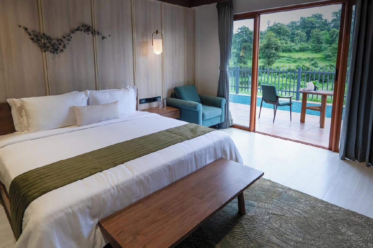 A hotel room with a large bed, a bench, a chair, a table, and a balcony with a view of greenery.