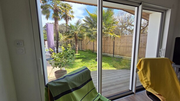 A room with a sliding glass door leading to a backyard with palm trees and a wooden deck. - Villa with private pool - 7 guests (Jard-sur-Mer)