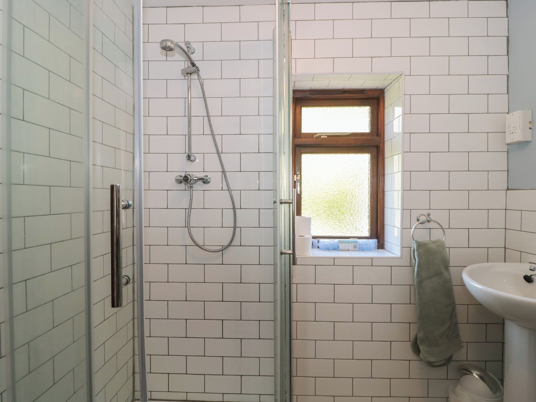 A bathroom with white tiled walls, a glass shower enclosure, a small window with a view, and a sink with a towel.