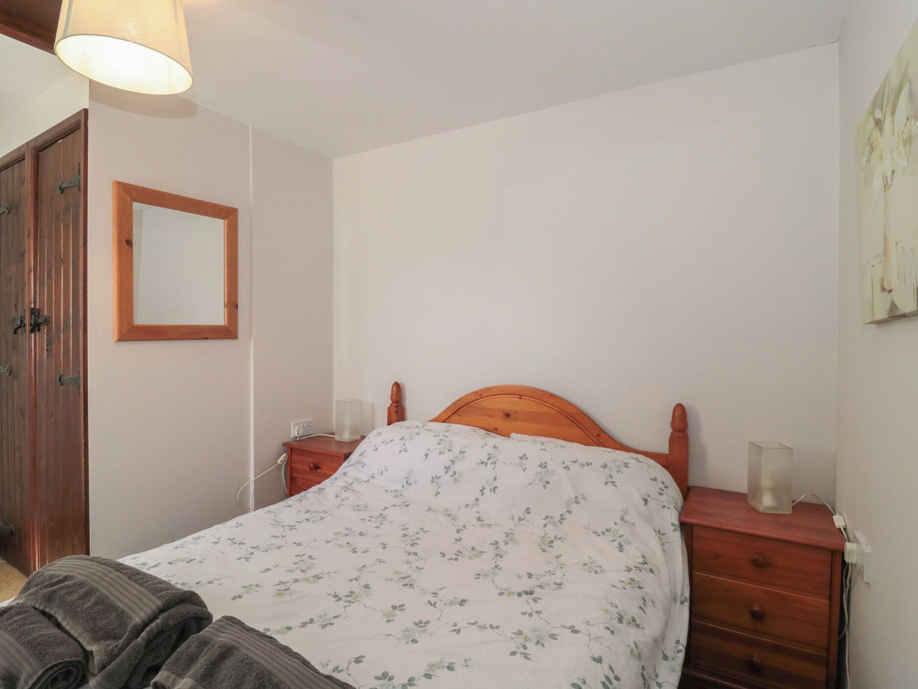 A bedroom with a wooden bed, a bedside table with a lamp, a mirror on the wall, and a wooden wardrobe.