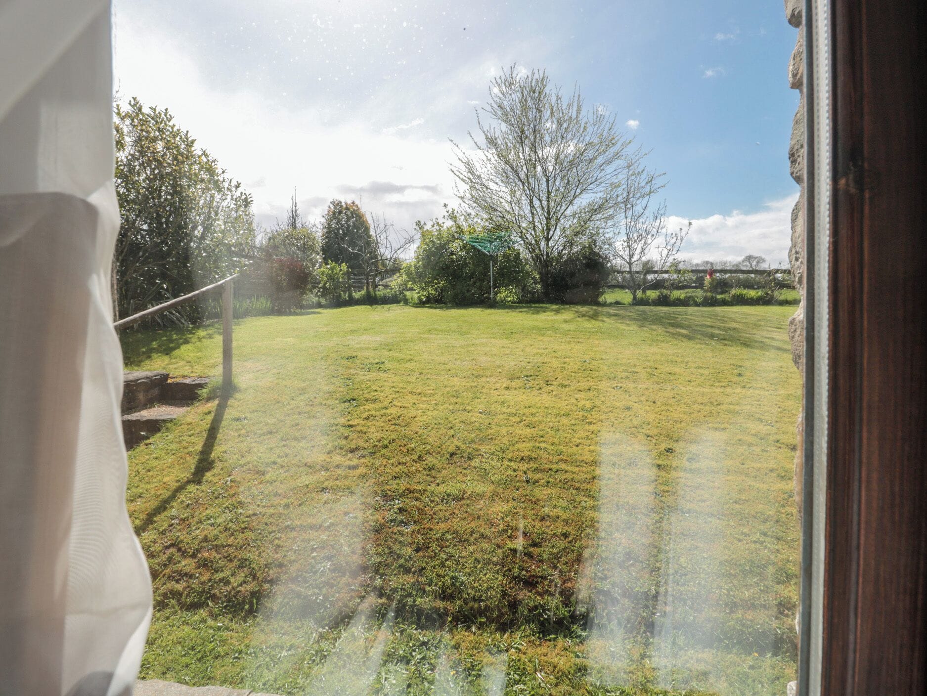 Vista a través de una ventana de un jardín con césped, árboles y una valla al fondo.