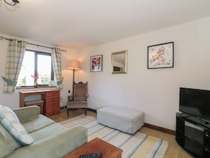 A living room with a sofa, a small table, a chair, a TV, a lamp, a window with curtains, and two framed pictures on the wall. - The Cowshed (Bristol)