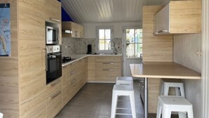 A compact kitchen with wooden cabinets, a microwave, a stove, and a sink. There are two white stools and a small table. - Comfortable house, pets welcome - 10 guests (Noirmoutier-en-l'Île)