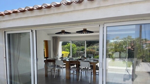 A modern house with a patio area featuring a dining table and chairs, large sliding glass doors, and a ceiling fan. - Comfortable house, pets welcome - 10 guests (Noirmoutier-en-l'Île)