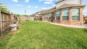 A residential backyard with a lawn, a wooden fence, and a brick house with large windows. - Cypress - Private Bedroom in Shared Home (Cypress)