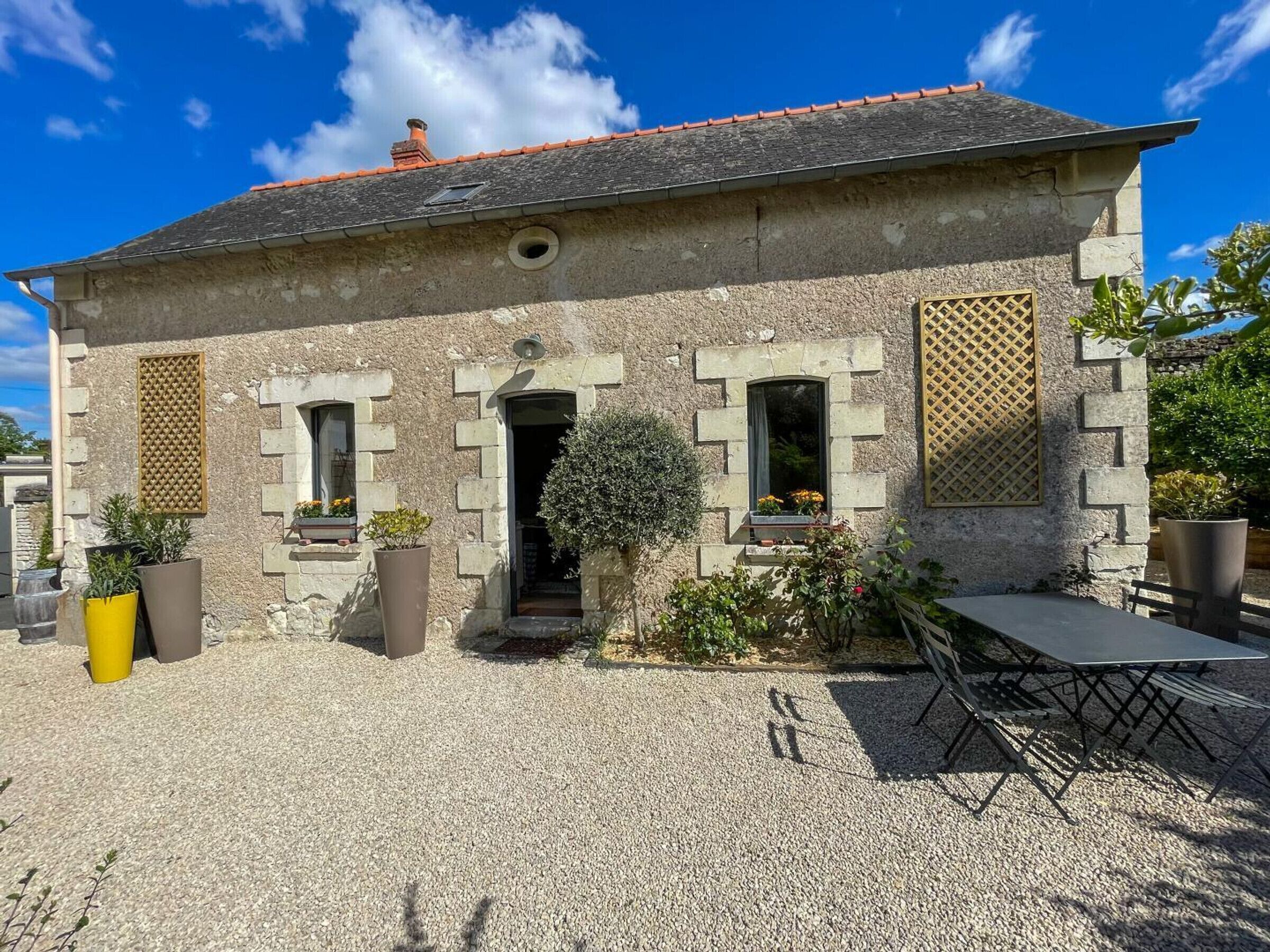 Ein Steinhaus mit Kiesweg, einer kleinen Terrasse mit Tisch und Stühlen und Topfpflanzen.