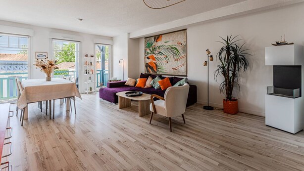 Een moderne woonkamer met een houten vloer, een eethoek met een tafel en stoelen, een bank met kussens, een groot schilderij aan de muur en een potplant.
