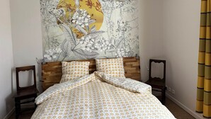 Un dormitorio con cama, cabecero de madera, dos mesitas de noche, un tapiz en la pared y una cortina amarilla.