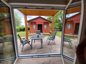 Une terrasse fermée par des baies vitrées, avec une table et des chaises, une petite maison en arrière-plan et une zone herbeuse.