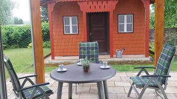 Ein überdachter Patio-Bereich mit Tisch und Stühlen, ein kleines rotes Gebäude im Hintergrund und Blick durch eine Glastür.