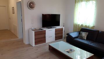 Ein Wohnzimmer mit einem Flachbildfernseher auf einer weißen Holzeinrichtung, einer schwarzen Ledersofa und einem Glastisch.