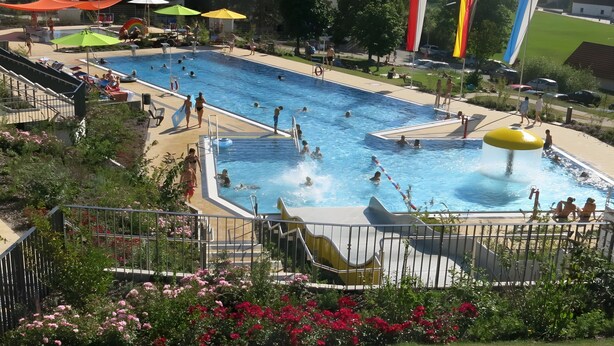 Ein Schwimmbecken mit mehreren Bahnen, einer Wasserrutsche und einem von Blumen gesäumten, eingezäunten Bereich.