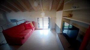 A room with a red sofa, a flat-screen TV, wooden beams on the ceiling, and a large window. - Attic Boutique May (Bergamo)