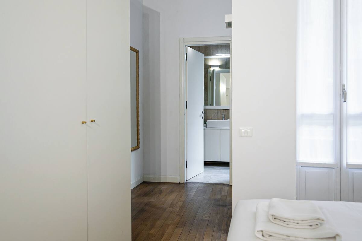 A hotel room with a wooden floor, a bed with a white comforter, a white wardrobe, and a bathroom visible through an open door.