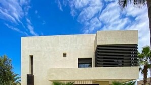 A modern two-story house with a flat roof, large windows, and a well-maintained lawn with palm trees.