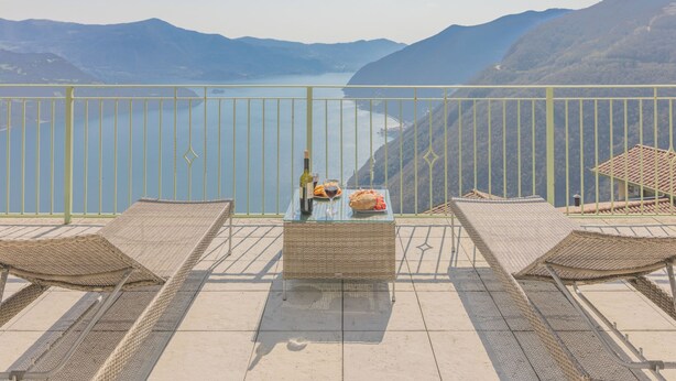 Varanda com mobiliário de vime, uma mesa com garrafa e copos, e vista para um lago e montanhas.