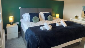 Una cama bien tendida con almohadas y una colcha, una lámpara de noche, una mesita con lámpara y un espejo en la pared.