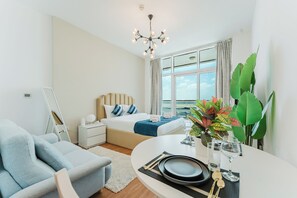 A modern hotel room with a bed, a desk with a vase, a chair, and a large window. - PVH - Al Jawhara - Charming Studio (Dubai)