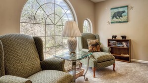 A room with large arched windows, a glass coffee table, two green upholstered chairs, a lamp, a bookshelf, and a wall-mounted bear illustration. - Amazing Gatlinburg Views - Briarcliff Subdivision, Hot Tub, Game Room, Sleeps 8 (Gatlinburg)