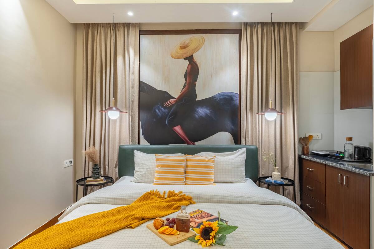 Quarto de hotel com cama, um quadro, uma mesinha com um livro, uma garrafa e um tabuleiro com fruta e um girassol.