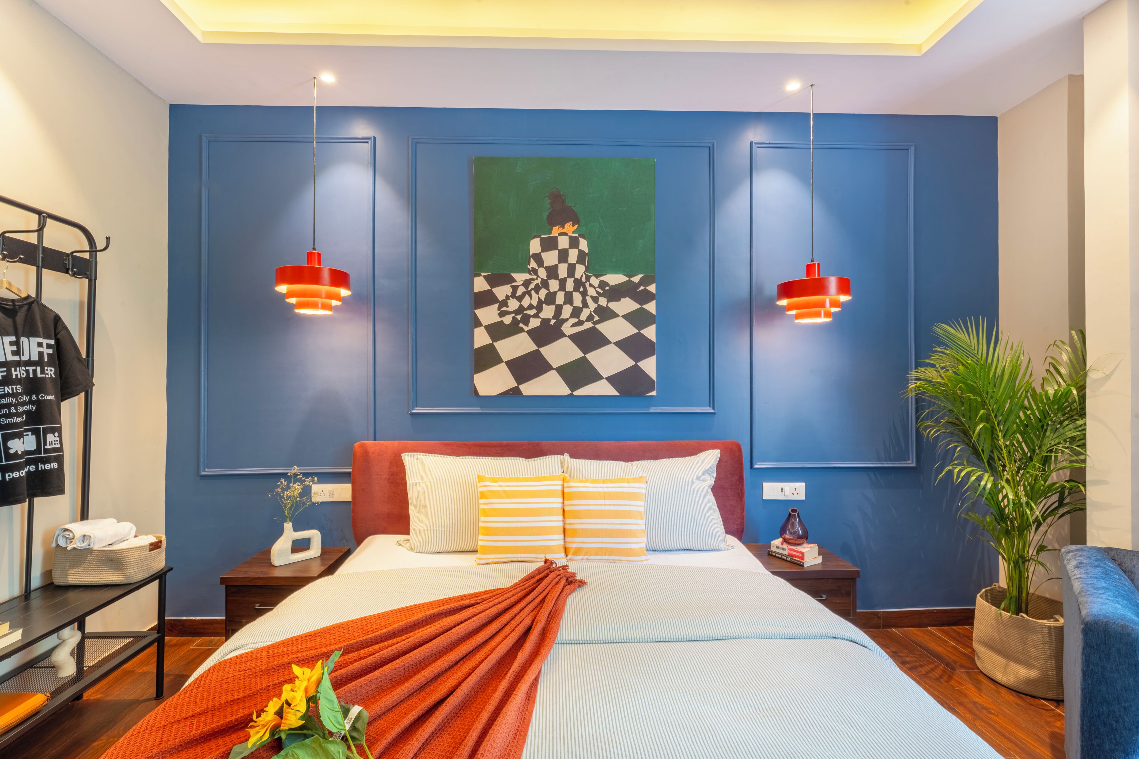 A hotel room with a bed, a nightstand, a desk, a chair, a plant, and artwork on the wall.