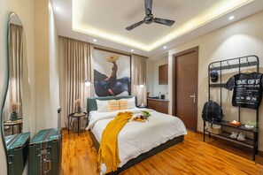 Una habitación de hotel moderna con una cama, un ventilador de techo, un espejo, un armario y una pequeña mesa con una bandeja de fruta y flores.