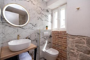 A bathroom with a marble wall, a white sink, a toilet, and a window.