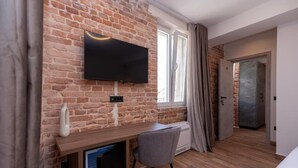 Ett rum med en tegelvägg, en väggmonterad TV, ett skrivbord med en stol, ett fönster med gardiner och en dörr som leder till ett annat rum.