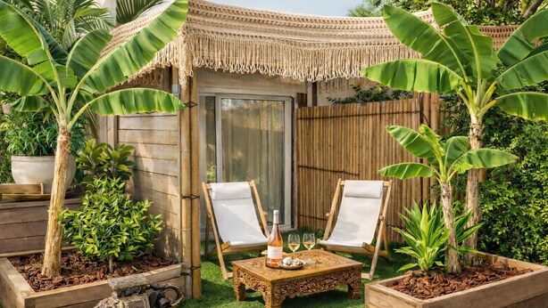 Un jardín tropical con un quiosco de madera, dos sillas blancas de lona, una mesita con una botella y vasos, y diversas plantas.