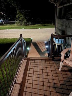 A wooden deck with a railing, a concrete pathway leading to a grassy area, and a chair on the right.