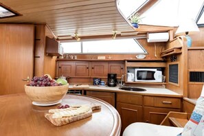 Cocina compacta con armarios de madera, microondas, lavabo y zona de comedor con un bol de fruta y queso.