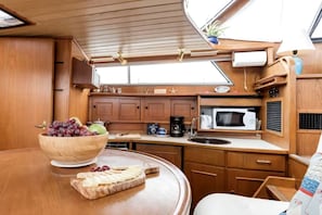 Cocina compacta con armarios de madera, microondas, lavabo y zona de comedor con un bol de fruta y queso.
