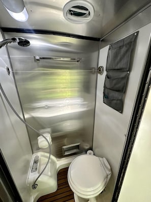 Une petite salle de bain aux finitions métalliques, équipée de toilettes, d’une douche et d’une poche de rangement.