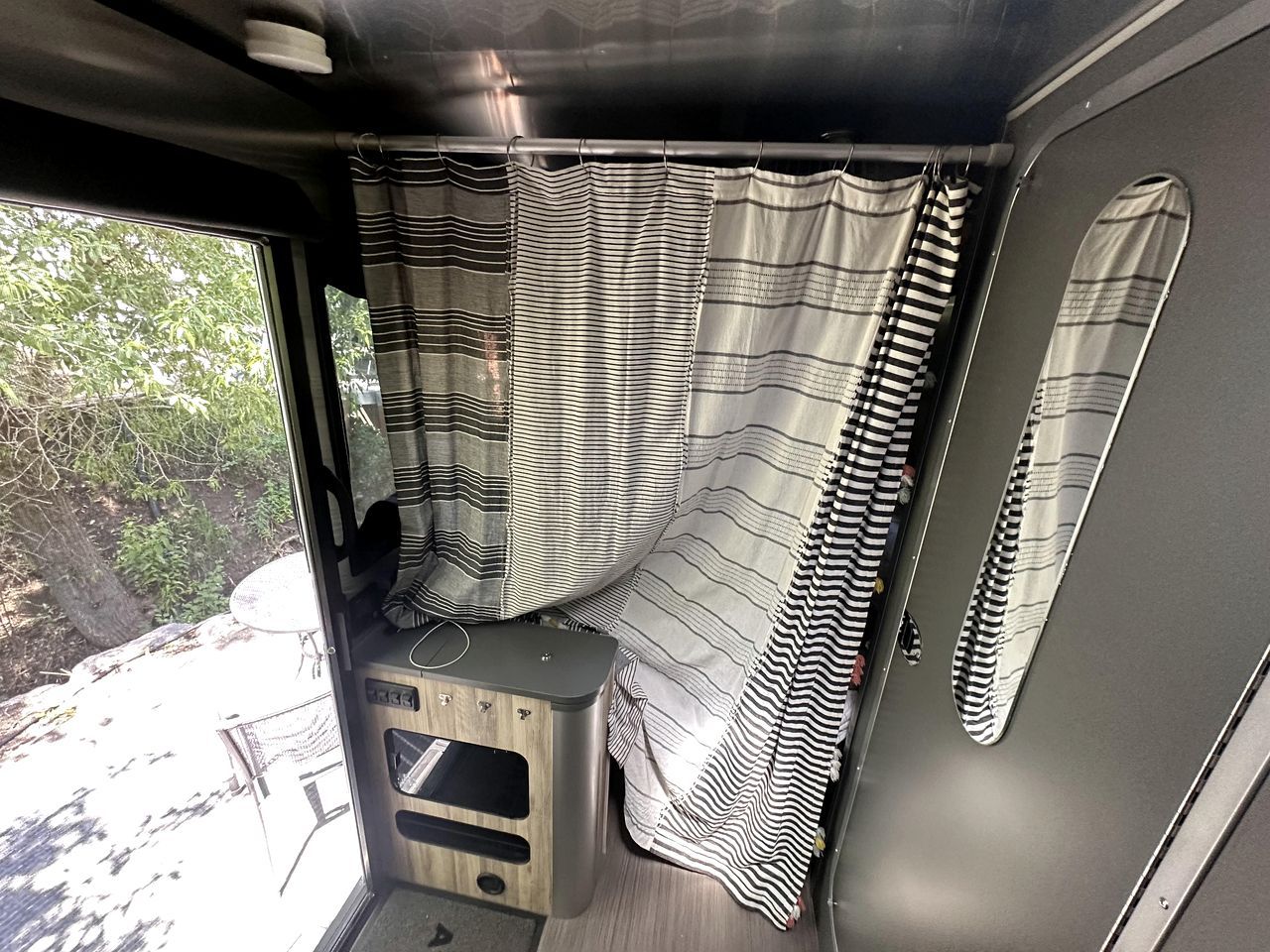 A van interior with a striped curtain, a small stool, and a window with a view of trees.