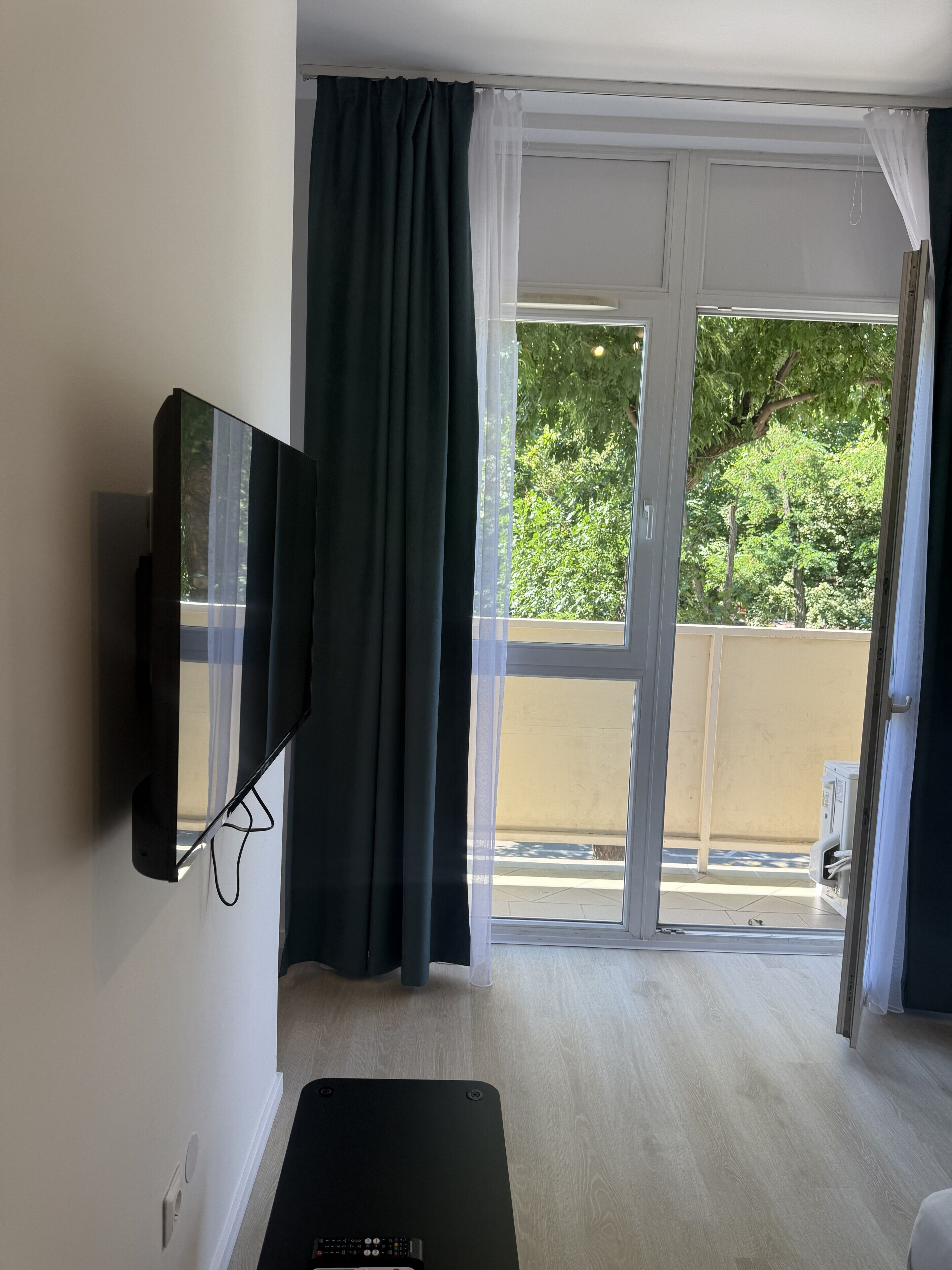Ein Zimmer mit Flachbildfernseher an der Wand, einer Glasschiebetür, die auf einen Außenbalkon führt, und Holzboden.