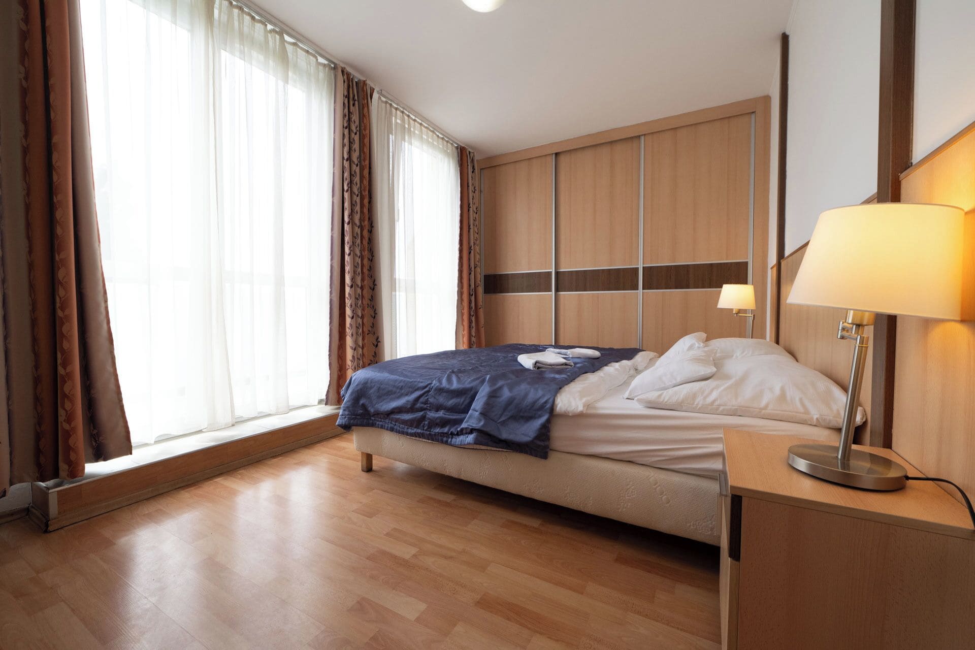 Une chambre d’hôtel avec un lit, une lampe de chevet, une armoire en bois et une fenêtre avec des rideaux.