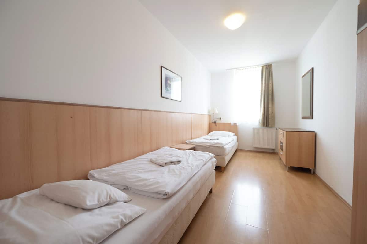 A hotel room with two single beds, a wooden headboard, a small bedside table, a radiator, a mirror, and a window with curtains.
