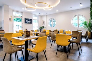 Un interior de restaurante moderno con sillas amarillas y marrones, mesas redondas y un techo con lámparas circulares.