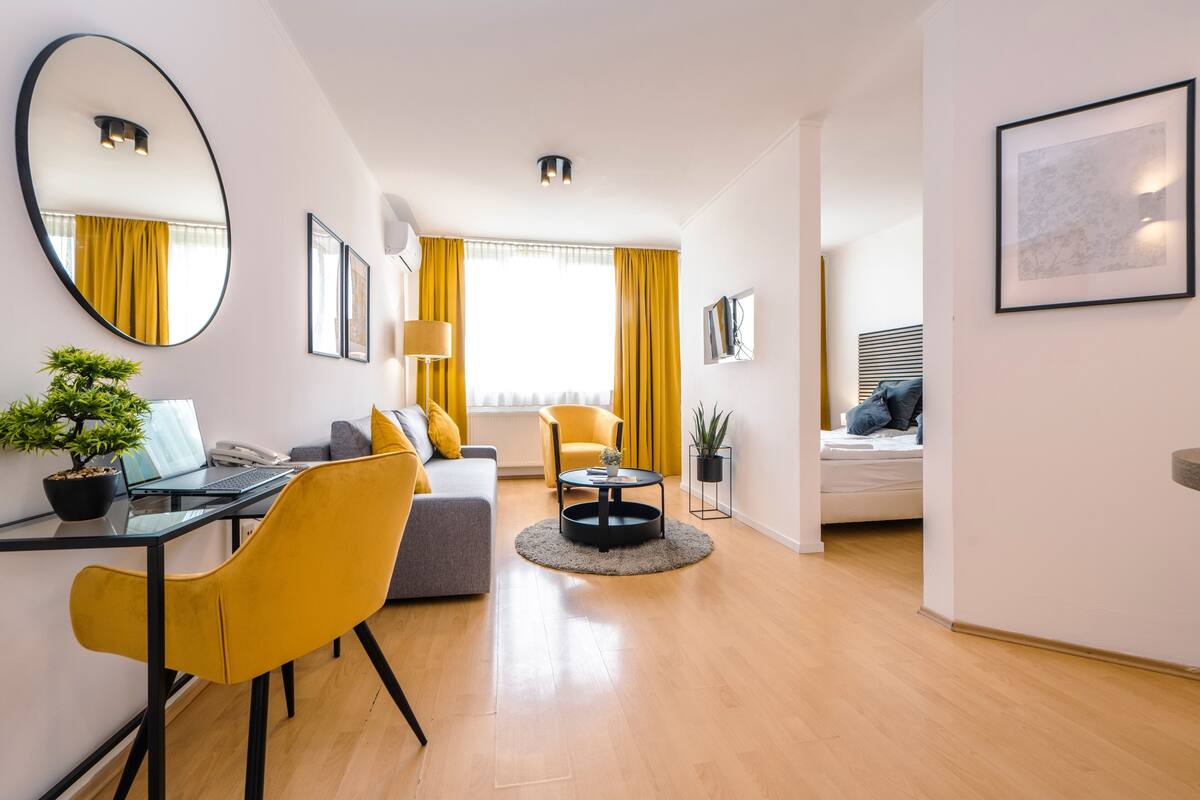 A modern living room with a sofa, yellow chairs, a round coffee table, a desk with a laptop, a potted plant, and a large mirror.