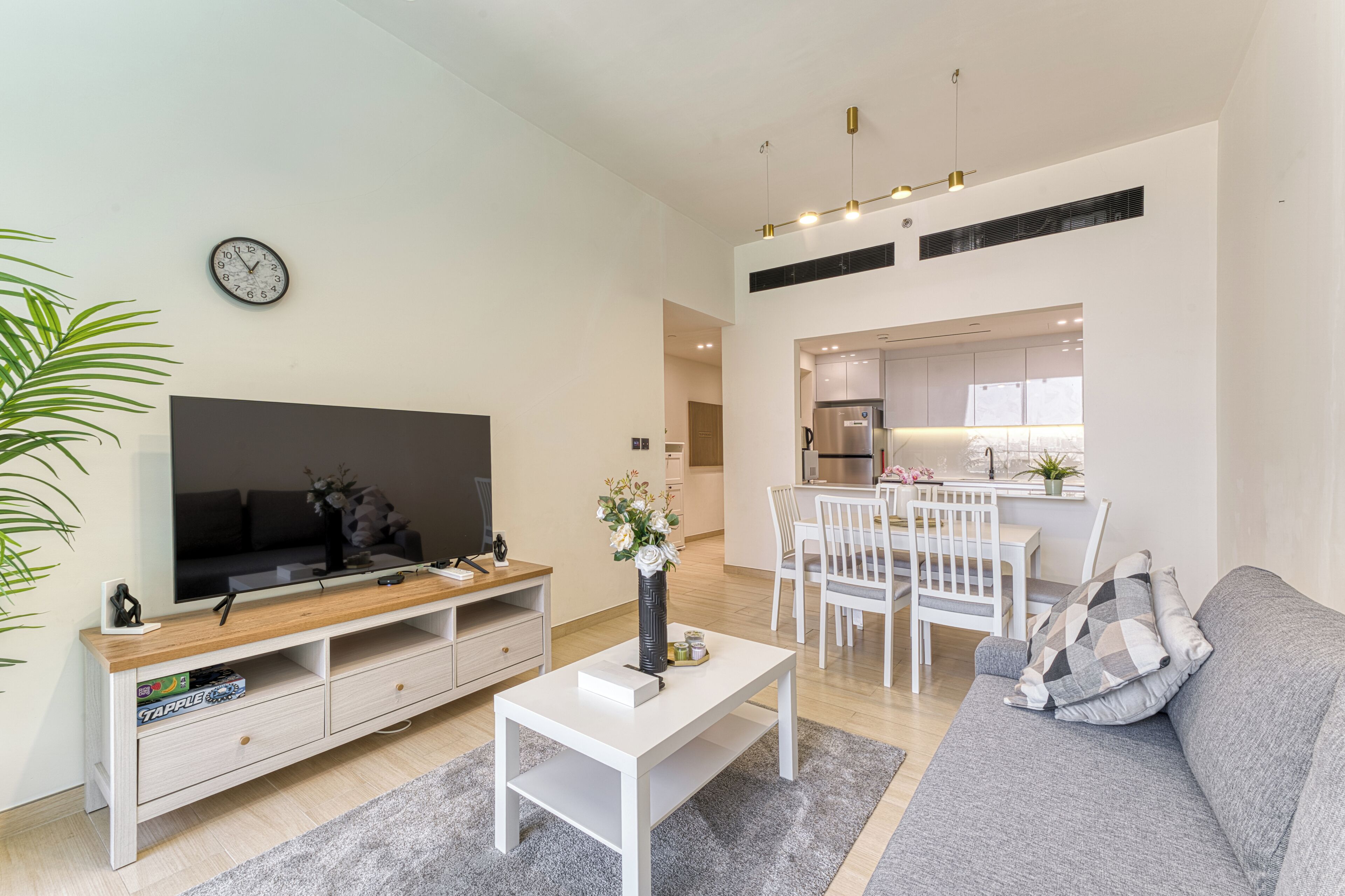 A modern living room with a flat-screen TV, a coffee table, a sofa, and a dining area with chairs and a kitchen in the background.