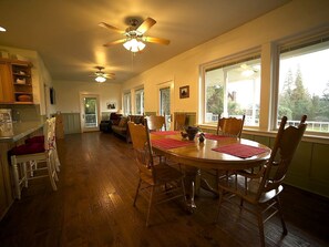 A spacious dining area with a wooden table and chairs, a ceiling fan, and large windows.