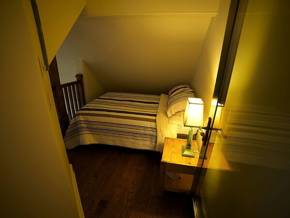 A small, dimly lit room with a single bed, a nightstand with a lamp, and a wooden door.