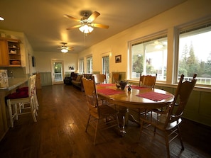 A spacious dining area with a wooden table and chairs, a ceiling fan, and large windows.