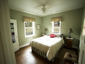 A bedroom with a bed, two windows with curtains, a ceiling fan, a nightstand, and a chair.