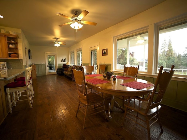 A spacious dining area with a wooden table and chairs, a ceiling fan, and large windows.